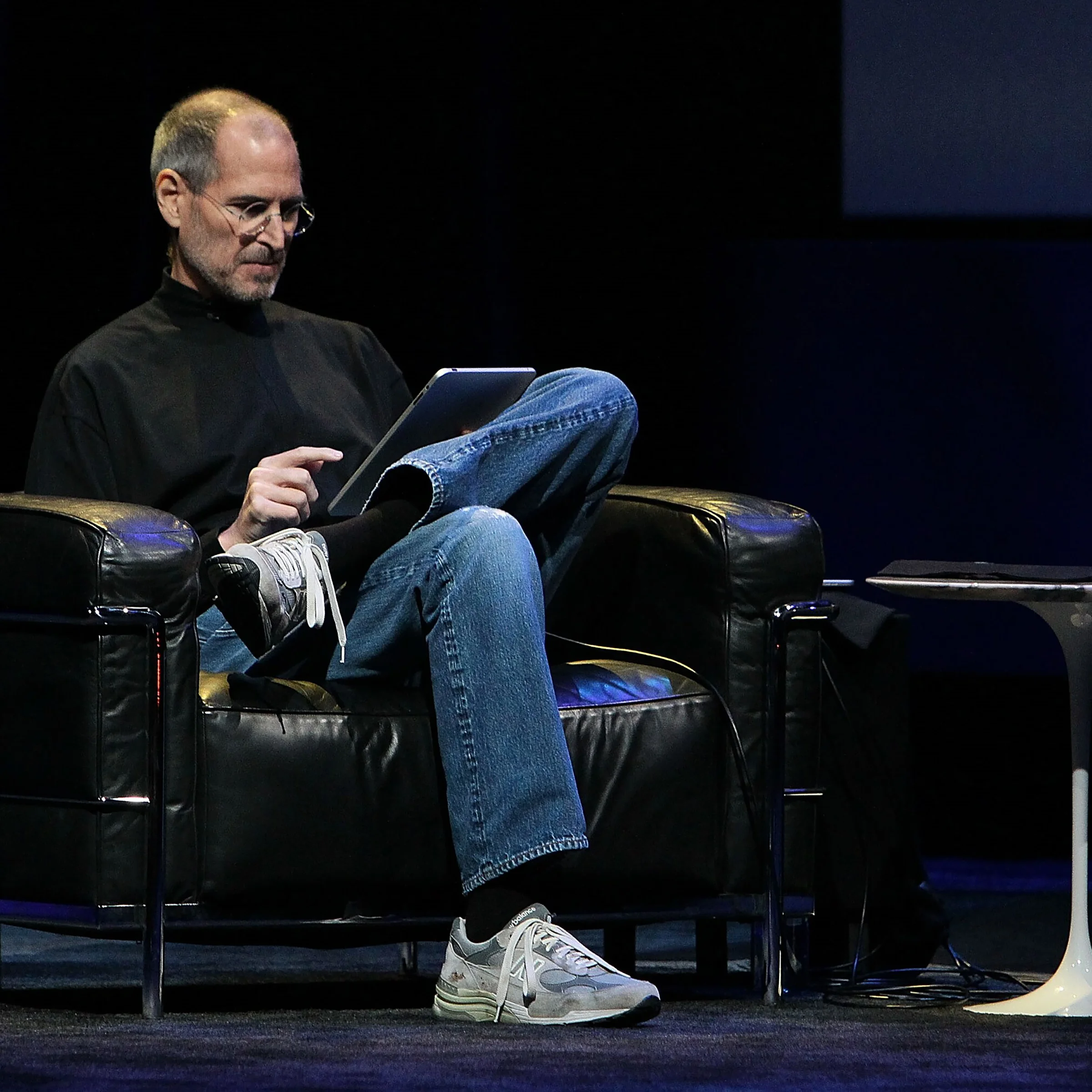 Steve Jobs sitting in a black armchair while introducing the first iPad in 2010, wearing grey New Balance shoes.